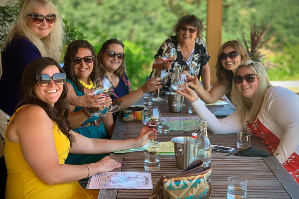 A group of women smiling and raising their wine glasses for a toast at an outdoor vineyard patio table.