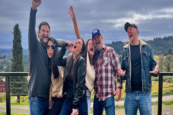 A lively group of friends cheering, laughing, and celebrating on a balcony overlooking scenic Willamette Valley vineyards.