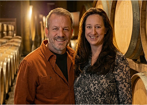Mark Treick and Helen McFarland, expert guides for Cellar Door Wine Tours, smiling together inside a Willamette Valley wine barrel room.